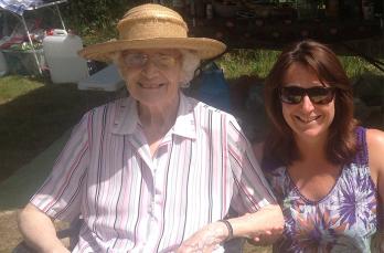 A smiling elderly woman wearing a summer hat sitting in a garden next to a middle aged smiling woman with brown hair
