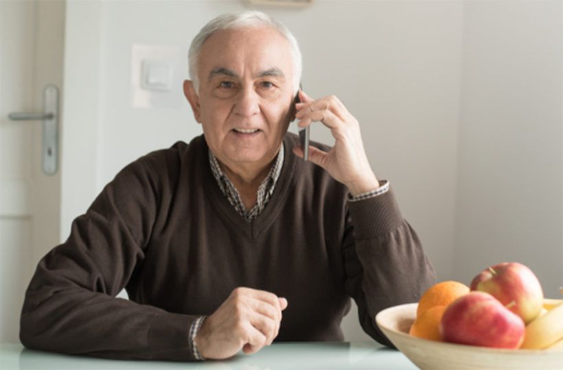 Older man sitting at a table, making a call using a mobile phone.
