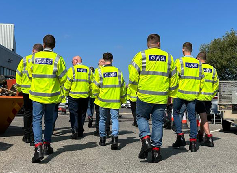 A line of SGN operatives, with their backs to the camera as they walk along in a row. The sky is a deep blue and their safety jackets are bright yellow.