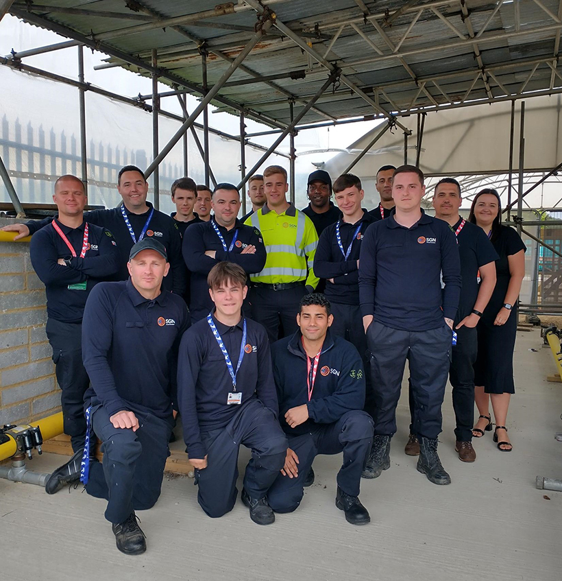 Group shot of 14 apprentices with an SGN team manager and East Sussex College tutor. They are standing in rows in the college's dedicated gas industry training facility