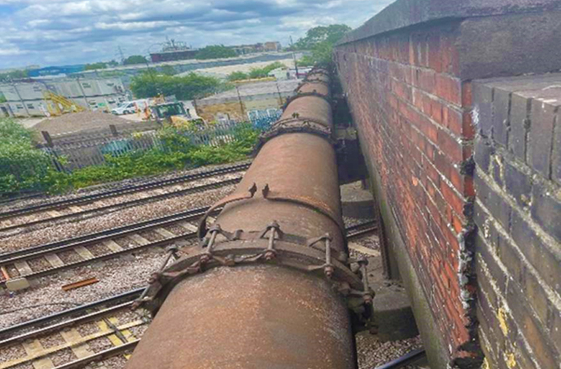 A metal pipeline attached to a bridge going over train lines