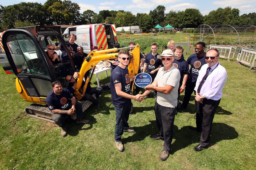 A group of people standing in a field beside a digger. Two at the front are shaking hands and holding a placard