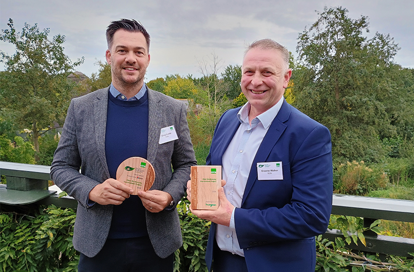 Two smiling men holding wooden awards against a nature background