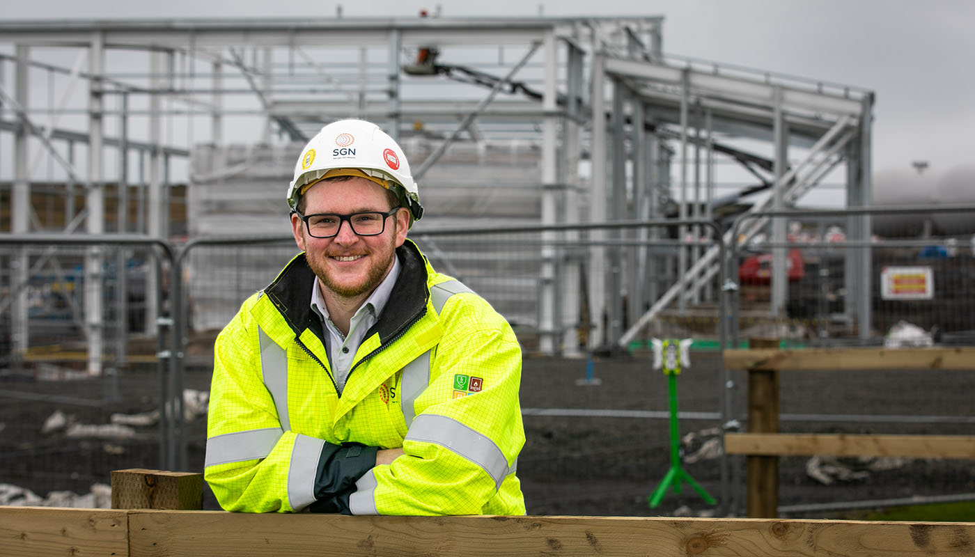 A man wearing PPE stands in front of an industrial site. 