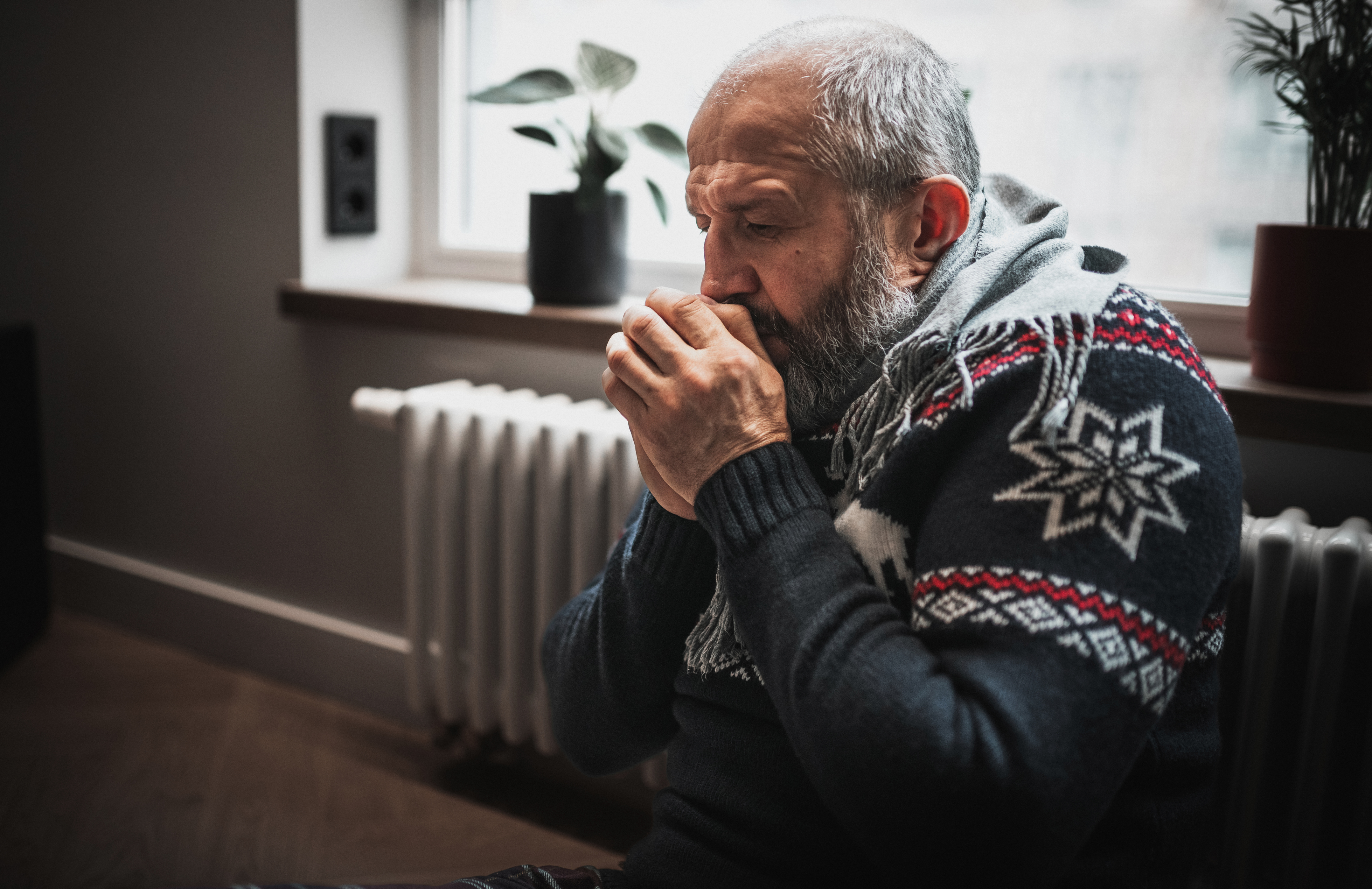 A man is sitting against a radiator, breathing into his cupped hands for warmth