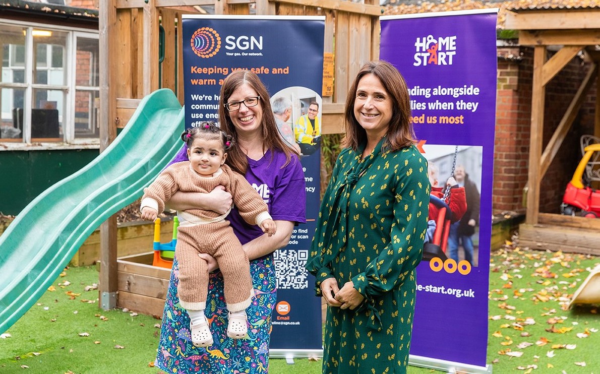 Colleagues from Home Start and SGN are standing in front of branded banners. The Home Start colleague is holding a toddler in her arms. 