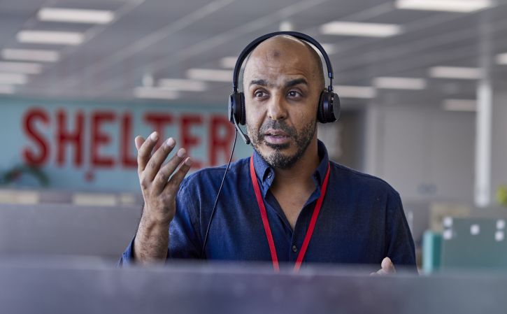 A Shelter helpline adviser is standing by his desk, wearing a telephone headset and gesturing forwards