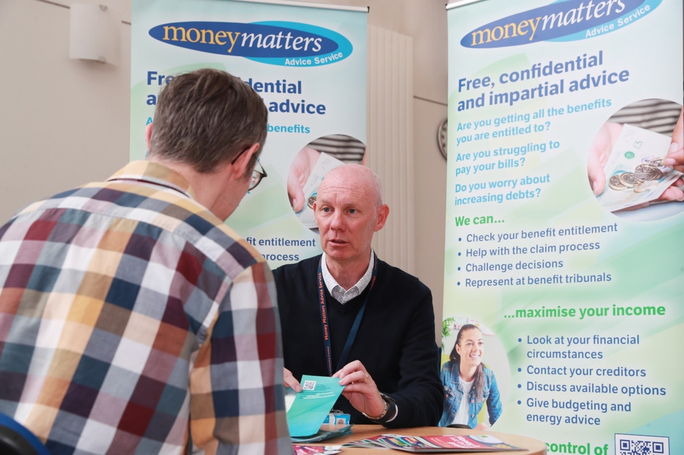 A Money Matters adviser is sitting at a table speaking with a South Lanarkshire Council resident. Behind him are two Money Matters information banners. 