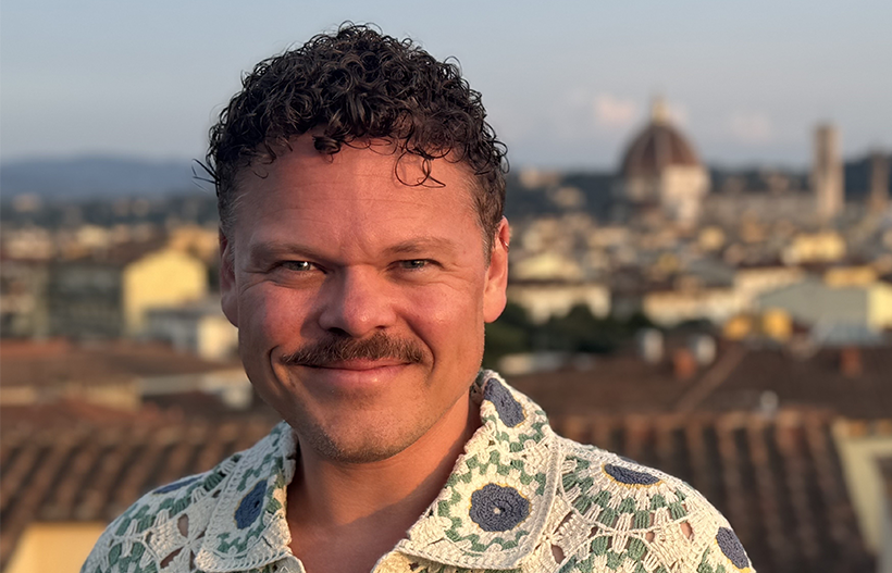 A smiling man with white skin, curly hair and moustache on the rooftop of a sunny European town