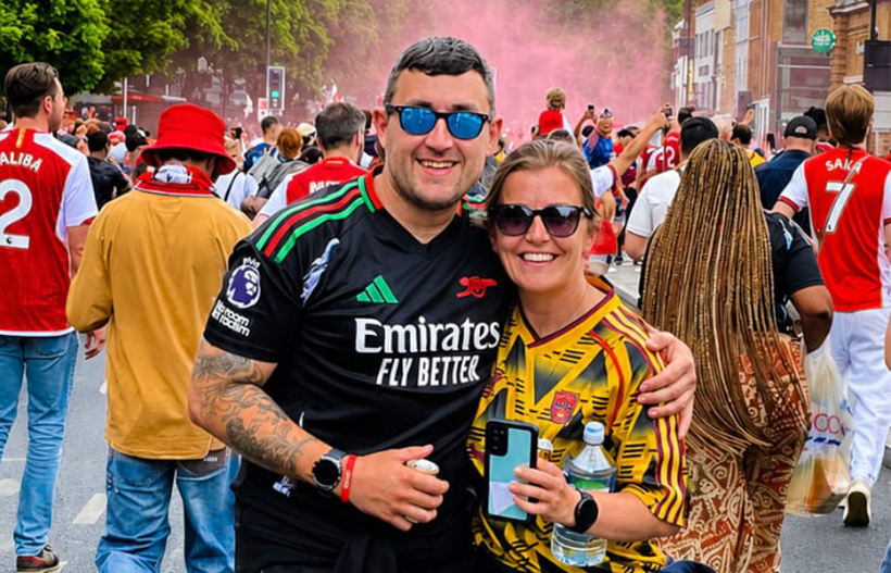 A man and woman in football shirts smiling at the camera surrounded by football fans