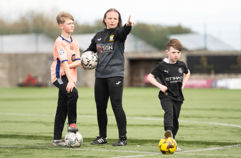An older teenage girl wearing a football tracksuit and holding a football. She is coaching two younger boys on a football pitch.