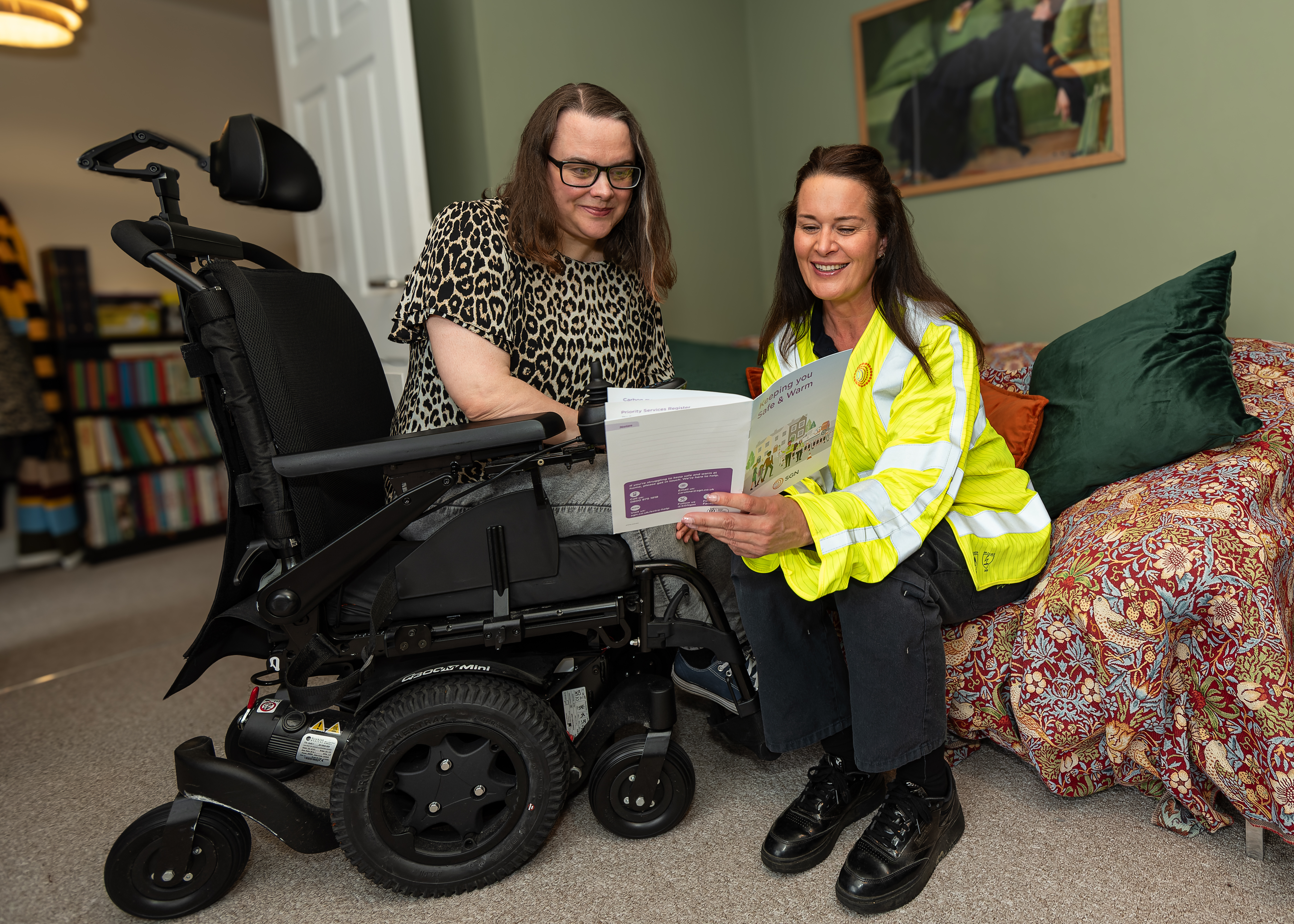 A member of the Safe and Warm team sat on a sofa holding a leaflet discussing its contents with a woman using a wheelchair