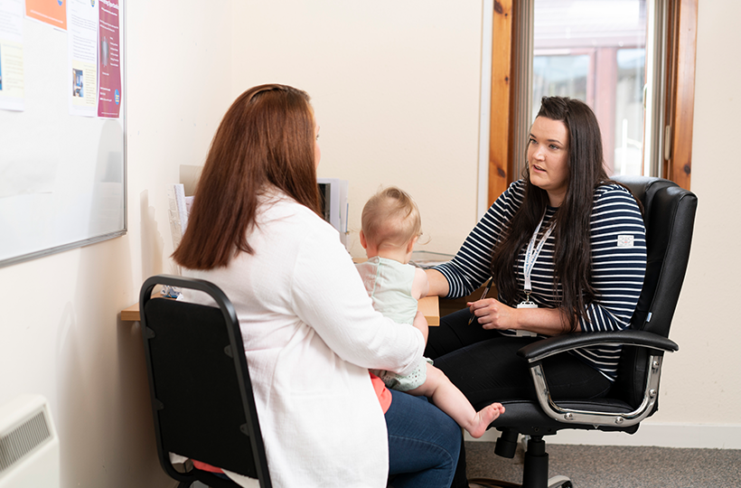 A person seated at a desk speaks with an adult holding a young child in a private office. A computer monitor, noticeboard and window are visible in the room.
