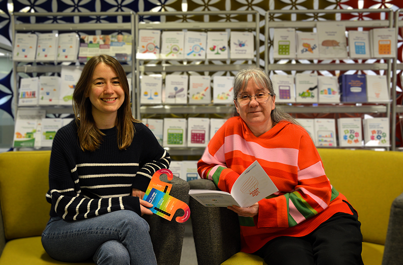 Two people seated on a yellow sofa in a library or learning space, holding brochures and printed materials, with shelves of educational booklets and guides visible in the background.