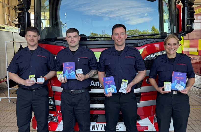 Four fire and rescue service staff stand in front of a fire engine holding home fire safety information packs and leaflets.