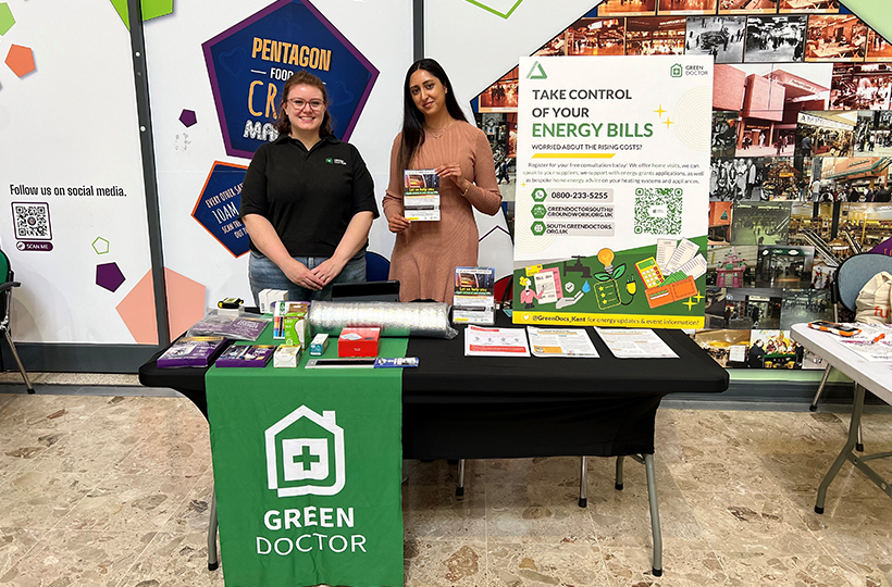 Two people stand behind a Green Doctor information stall with energy advice leaflets and a sign promoting support for managing energy bills.