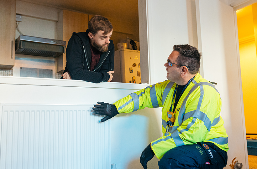 A worker wearing a high-visibility yellow jacket and gloves kneels beside a radiator in a flat, speaking with a resident who is leaning through a serving hatch from the kitchen. Kitchen units, an extractor hood and a fridge are visible in the background. The image shows a home visit related to heating or energy-efficiency advice.