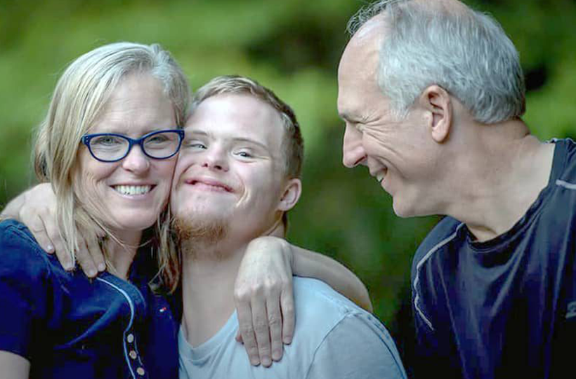 The image highlights a close, supportive interaction between the three individuals. Three people sit closely together outdoors with a blurred green background. The person in the centre, wearing a light grey T-shirt, has an arm around the shoulders of the person on the left, who is wearing blue-framed glasses and a dark blue top. The person on the right, wearing a dark navy shirt, leans towards them. 