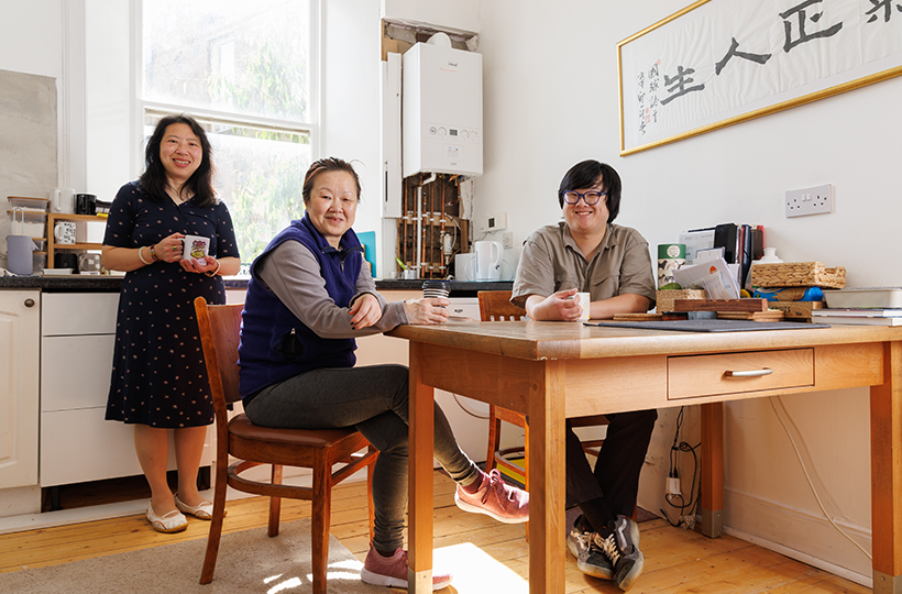 Three people are gathered in a bright office kitchen and meeting space. Two people sit at a wooden table holding drinks, while another person stands nearby holding a mug. The room has large windows, a kitchenette, a boiler mounted on the wall, and framed calligraphy artwork. Papers, folders and refreshments are arranged on the table and surrounding surfaces.