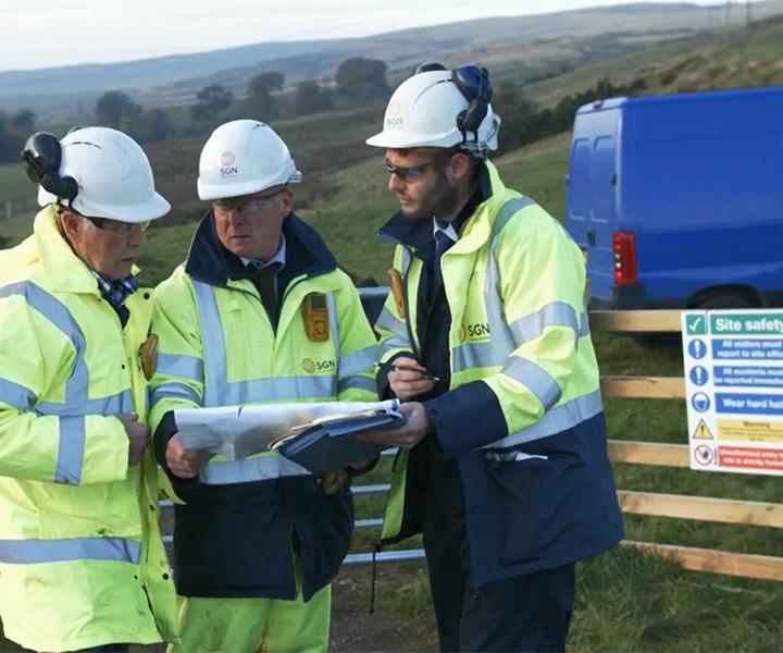 SGN colleagues standing with a customer in a field and are looking at a map.  The three people are wearing high-viz jackets and hard hats. 