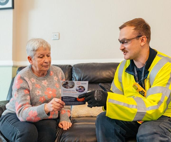 An engineer wearing PPE sits on a sofa next to an older woman. He is showing her a leaflet.