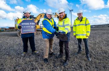 Four people wearing PPE standing in front of a digger machine. Two people holding a spade filled with dirt in the centre. One holds sign reading H100 Fife - it all starts here.