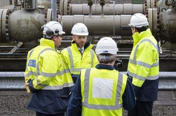 Four engineers wearing high-vis in discussion in front of pipeline behind