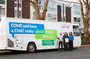 Three people stood in front of a converted bus - on its side: "Come and have a chat today about your health"