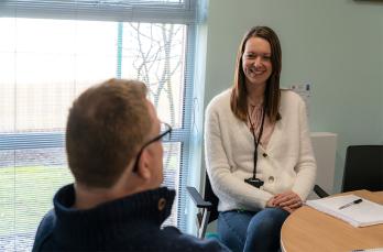 A smiling woman in an office wearing a lanyard speaking to a man